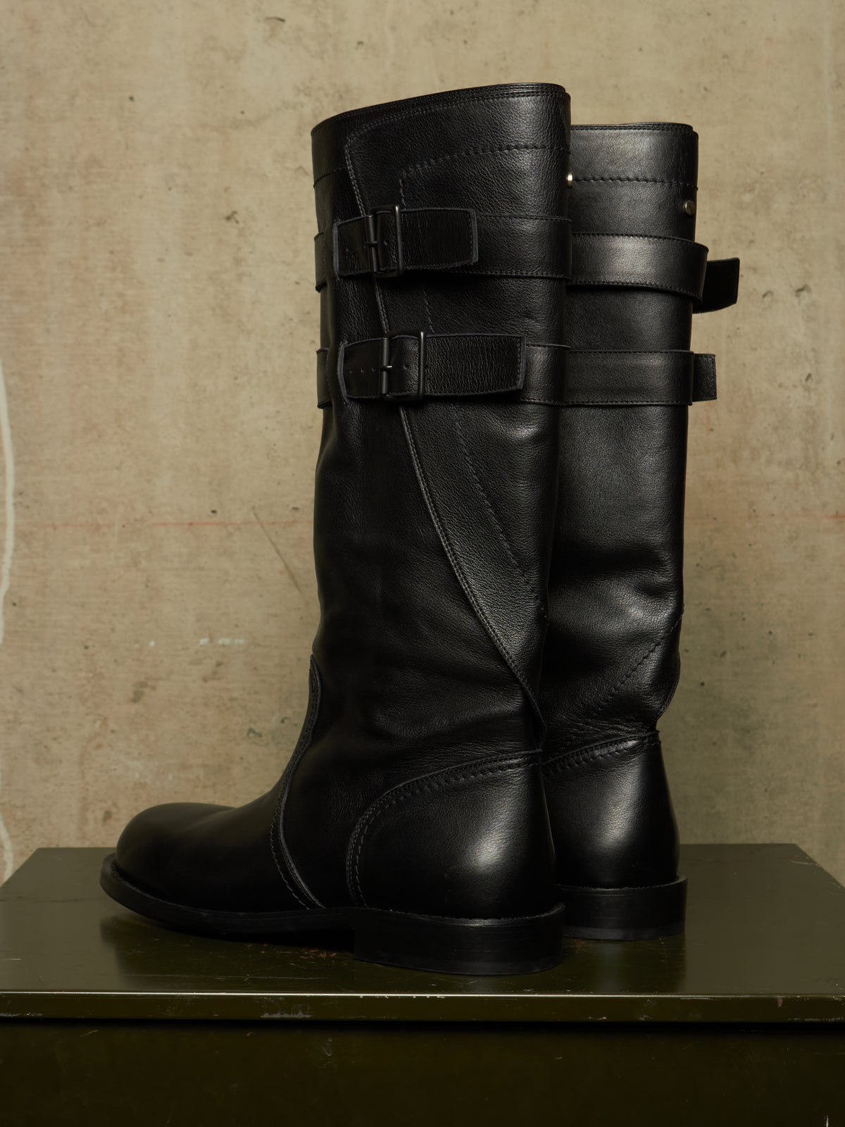 A pair of black leather knee-high boots with multiple decorative buckle straps, displayed on a dark green surface against a textured beige wall.
