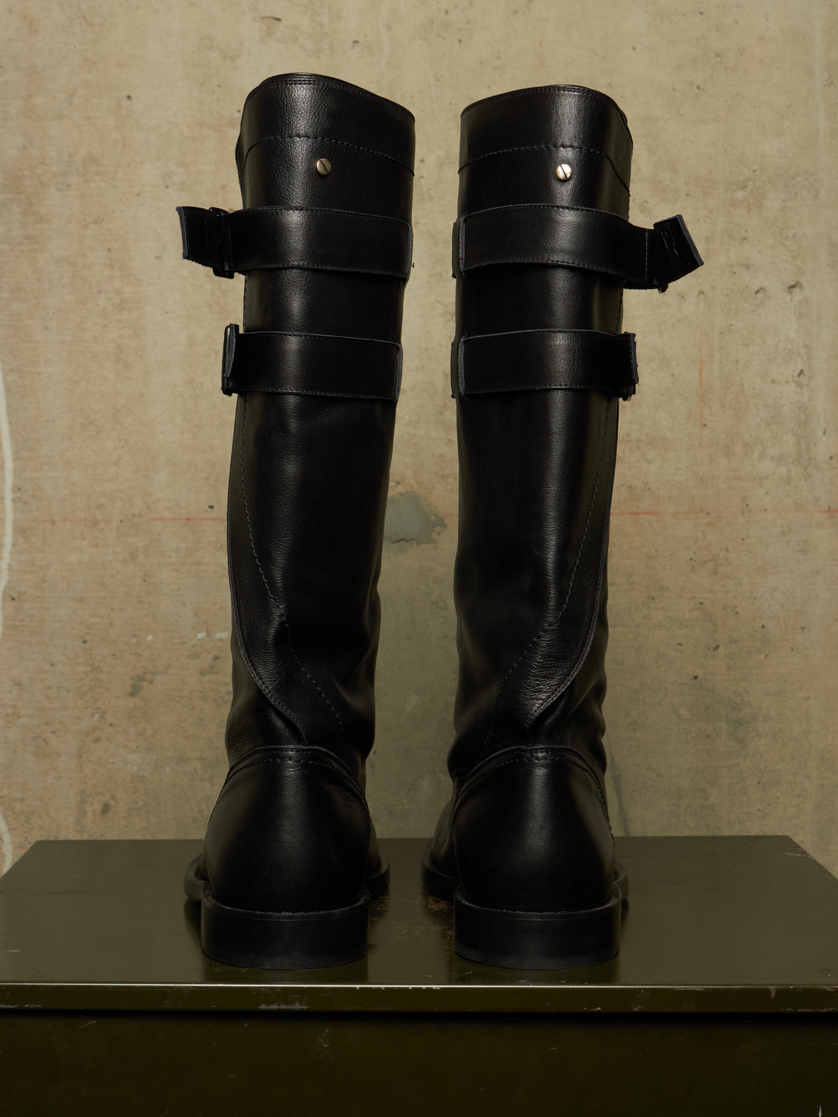 A pair of tall black leather boots with wide strap details and buckle closures, shown from the back on a dark surface against a textured beige wall.