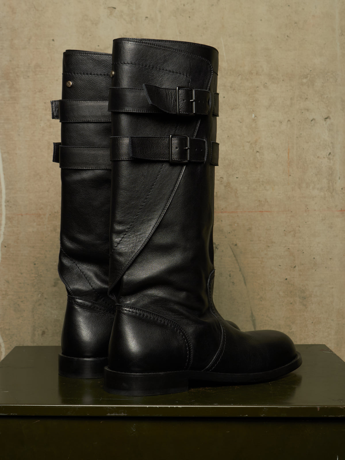 A pair of tall black leather riding boots with multiple decorative strap and buckle details on the shaft, displayed on a dark green surface against a textured beige wall.