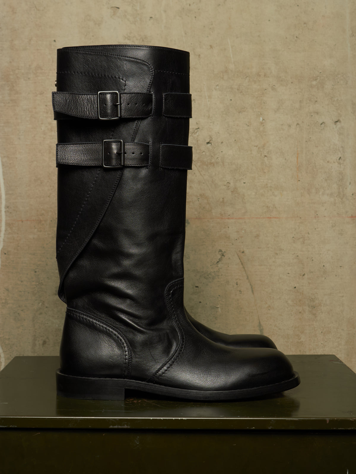 A tall black leather riding boot with two decorative buckle straps around the shaft, displayed on a dark surface against a textured beige wall.