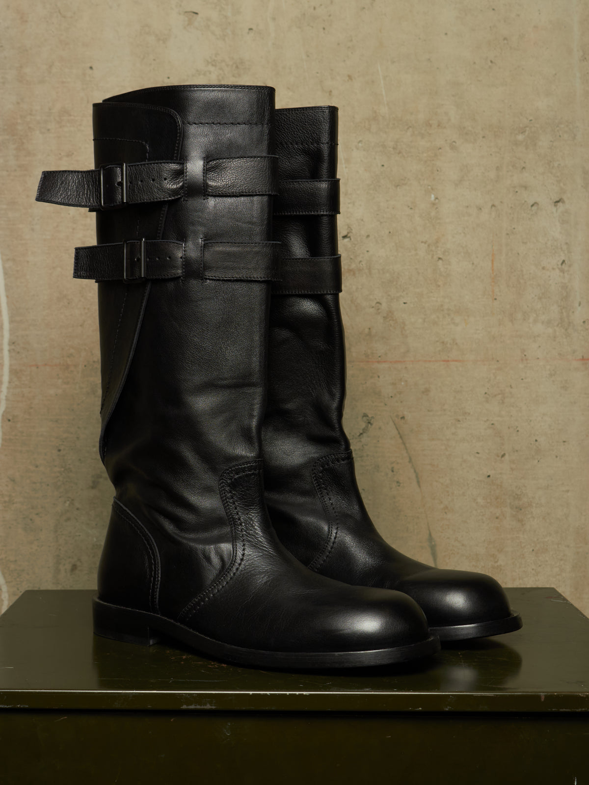 A pair of tall black leather boots with multiple strap details on the shaft, displayed on a dark green surface against a textured beige wall.