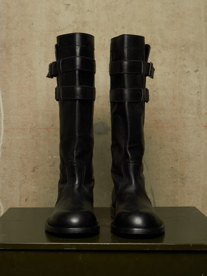 A pair of tall black leather boots with multiple strap details around the upper shaft, displayed side-by-side on a dark green surface against a textured beige wall.