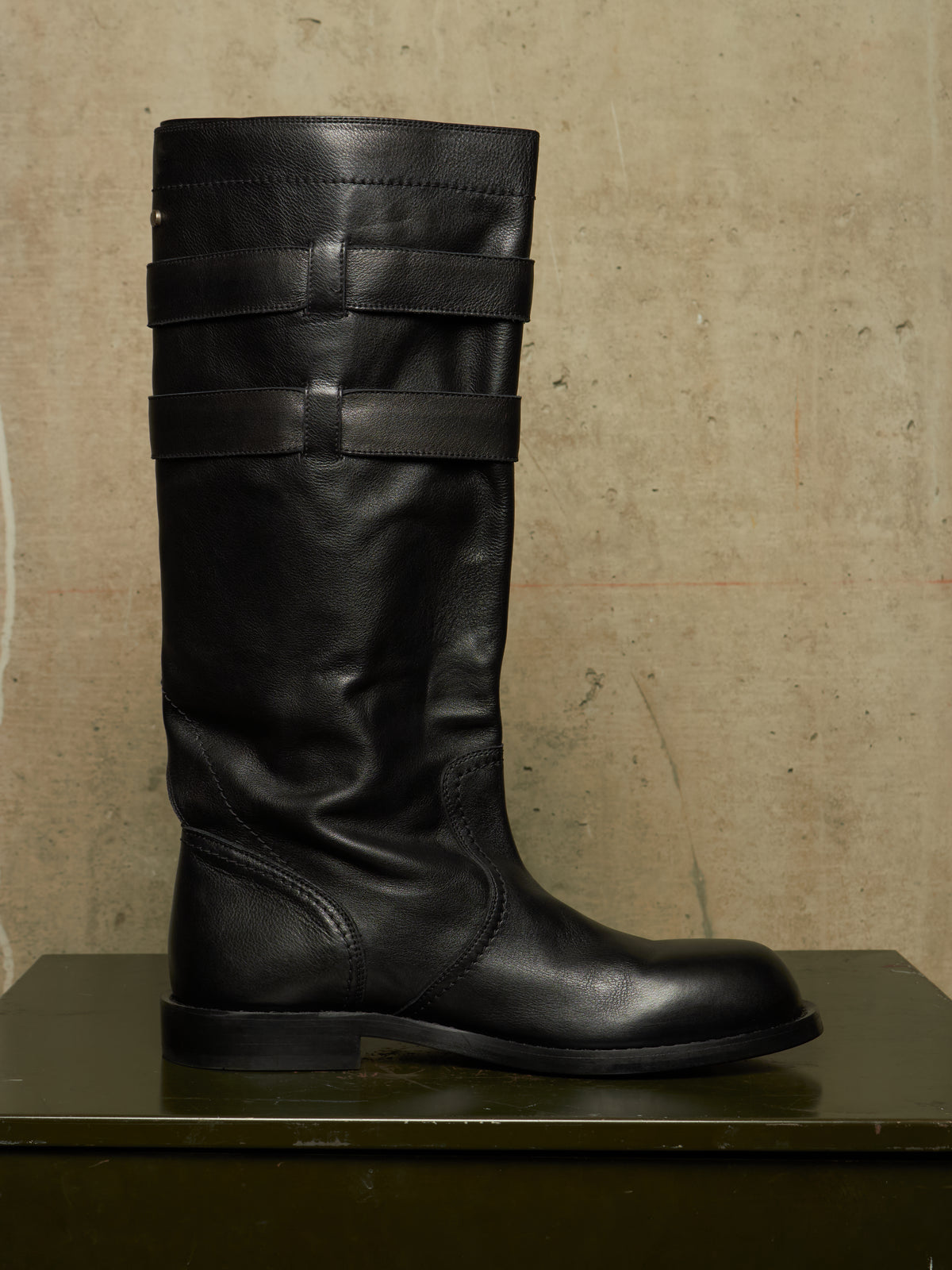 A single black leather knee-high boot with three horizontal strap details near the top, displayed on a dark green surface against a textured beige wall.