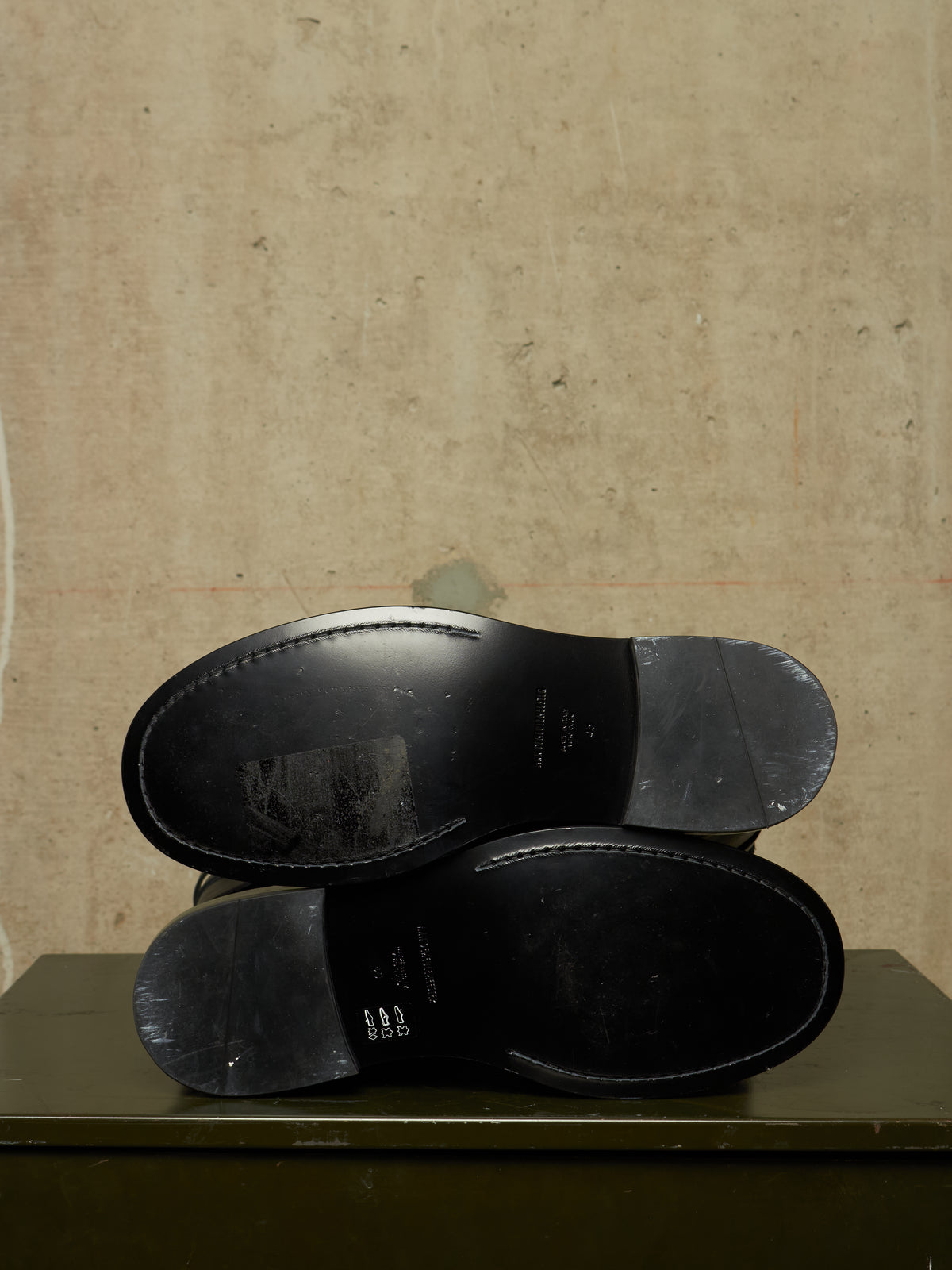 Two black leather shoe soles stacked vertically, showing the underside and heel caps, set against a textured beige wall on a dark green surface.