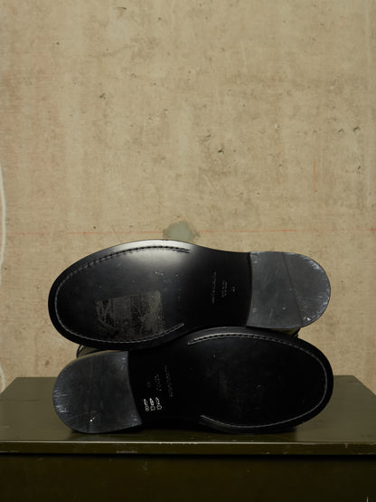 Two black leather shoe soles stacked vertically, showing the underside and heel caps, set against a textured beige wall on a dark green surface.