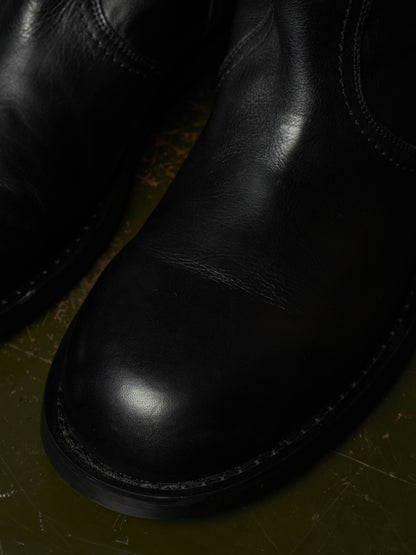Close-up of a black leather boot with a rounded toe and visible stitching, resting on a dark textured surface.