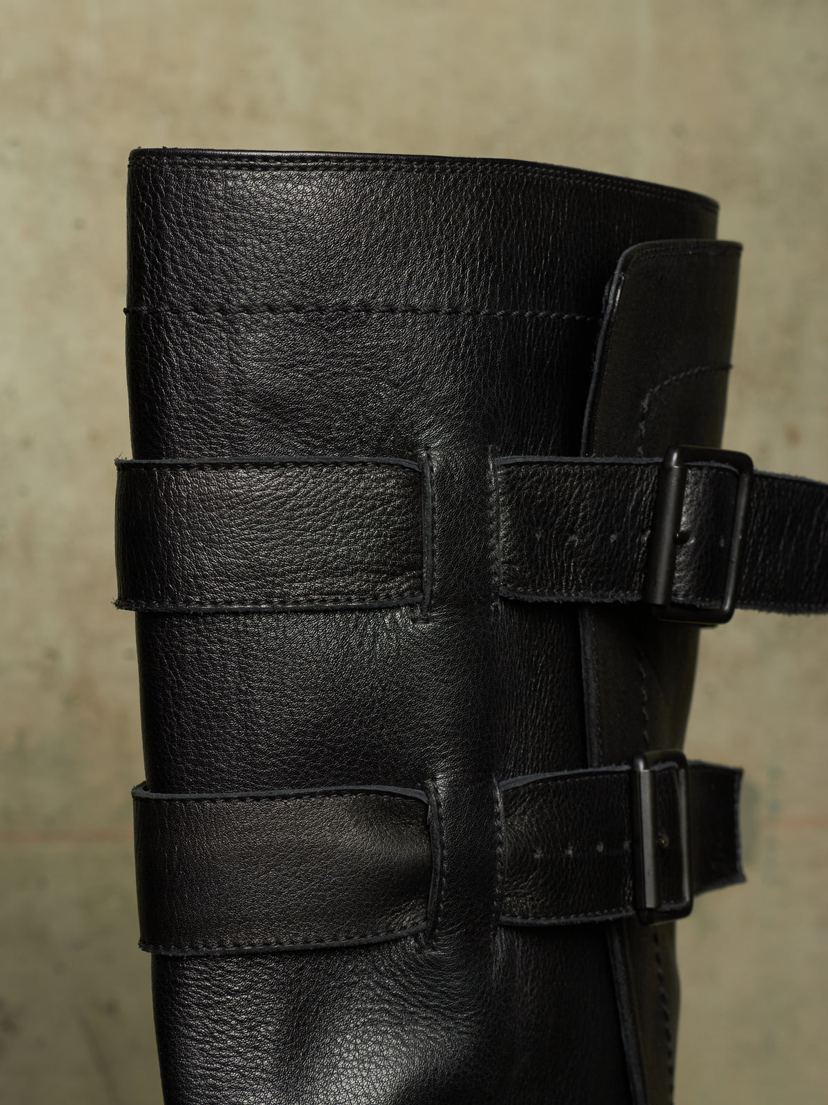 Close-up of a black leather boot with multiple wide decorative straps and metal buckle closures, shown against a neutral textured beige background.
