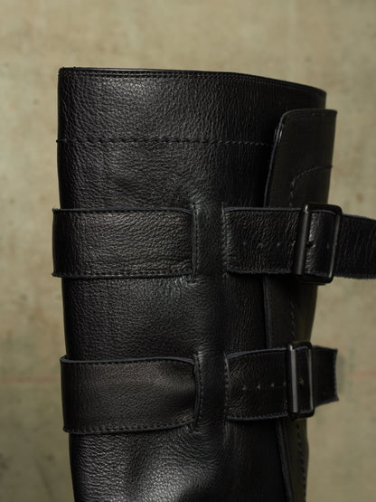 Close-up of a black leather boot with multiple wide decorative straps and metal buckle closures, shown against a neutral textured beige background.