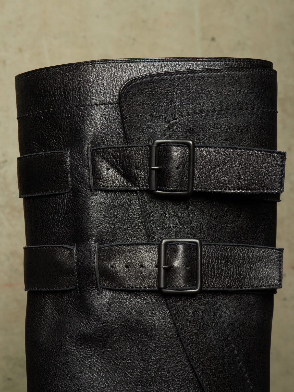 Close-up of a black leather boot shaft featuring two wide decorative straps with square metal buckles, set against a neutral textured background.