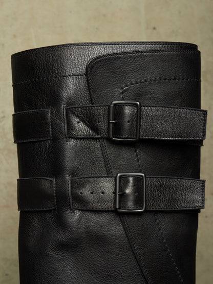 Close-up of a black leather boot shaft featuring two wide decorative straps with square metal buckles, set against a neutral textured background.