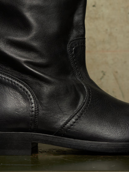 Close-up of a black leather boot showing the side stitching, textured leather surface, and low stacked heel against a neutral concrete background.