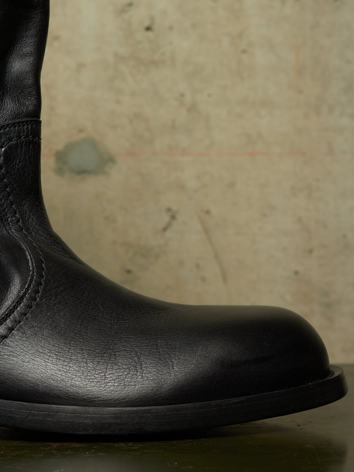 Close-up of a black leather boot showing the side profile and toe, set against a textured beige concrete background.