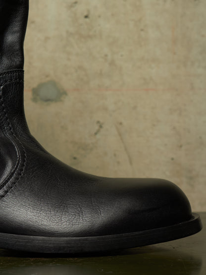 Close-up of a black leather boot showing the side profile and toe, set against a textured beige concrete background.