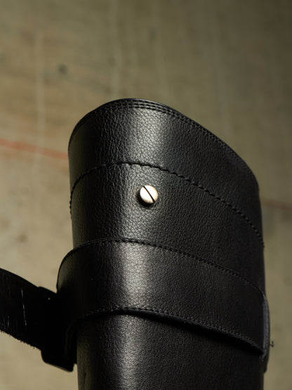Close-up of a black leather boot or shoe cuff with a strap and a small silver rivet, photographed against a textured gray background.