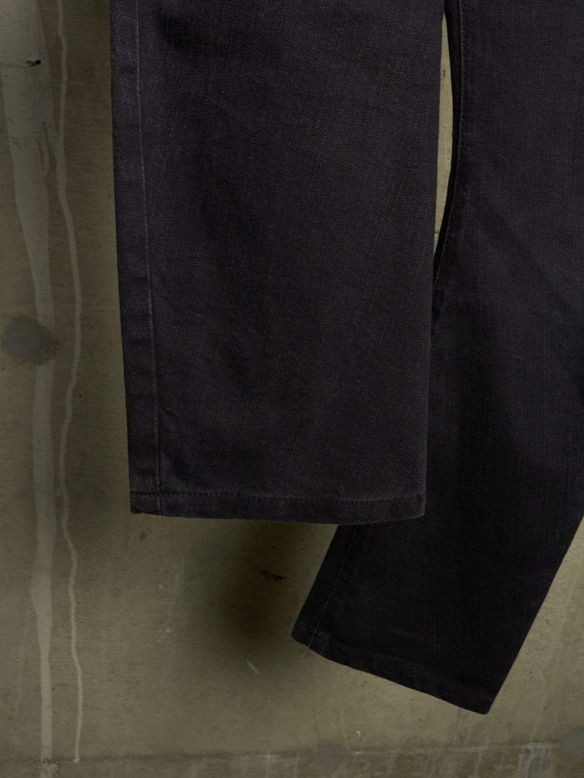 Close-up of a dark charcoal gray or black denim garment, showing the fabric texture and hem, set against a textured concrete background.