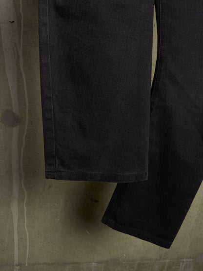 Close-up of a dark charcoal gray or black denim garment, showing the fabric texture and hem, set against a textured concrete background.