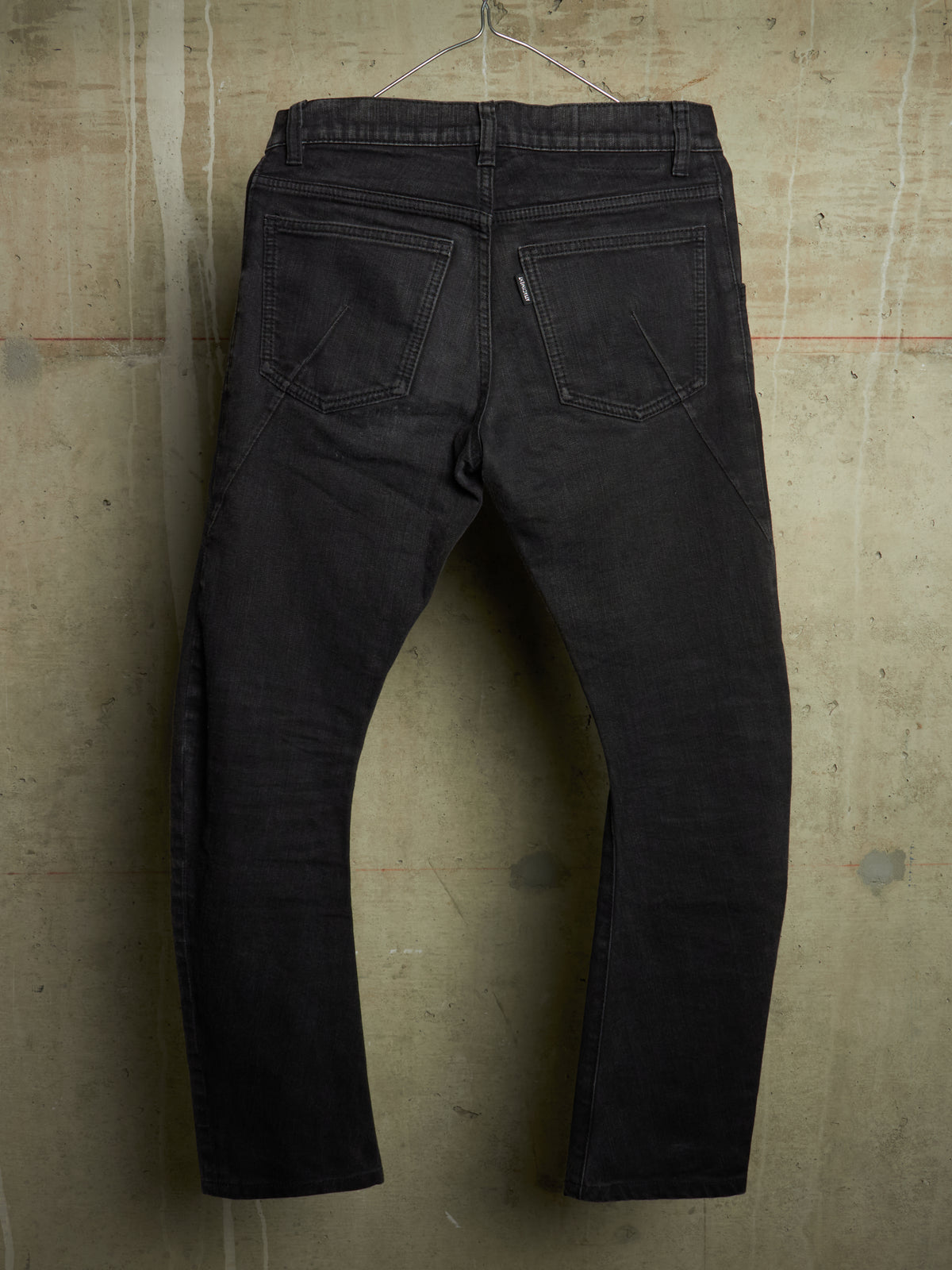 Black denim jeans shown from the back, hanging against a textured concrete wall.