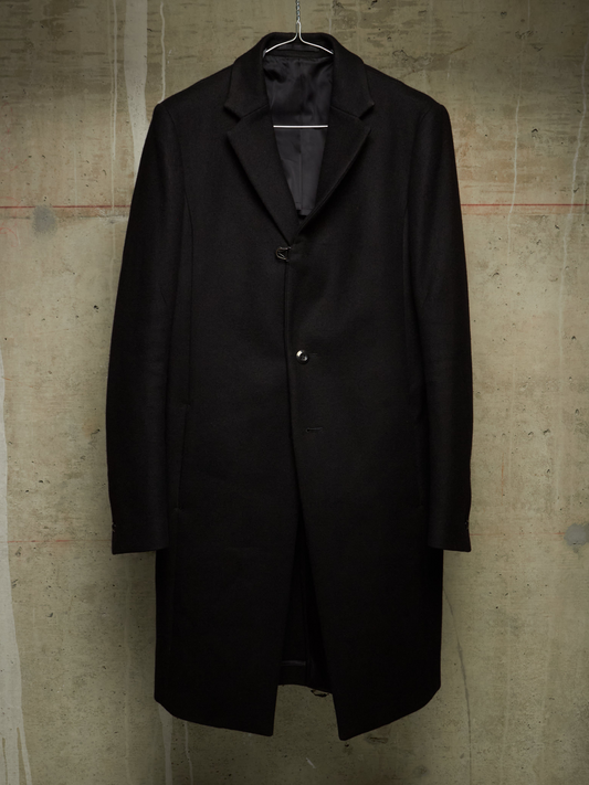 A black wool overcoat hanging on a metal hanger against a textured concrete wall.