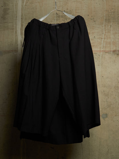 Black wide-leg pleated trousers hanging on a white plastic hanger against a textured concrete wall.