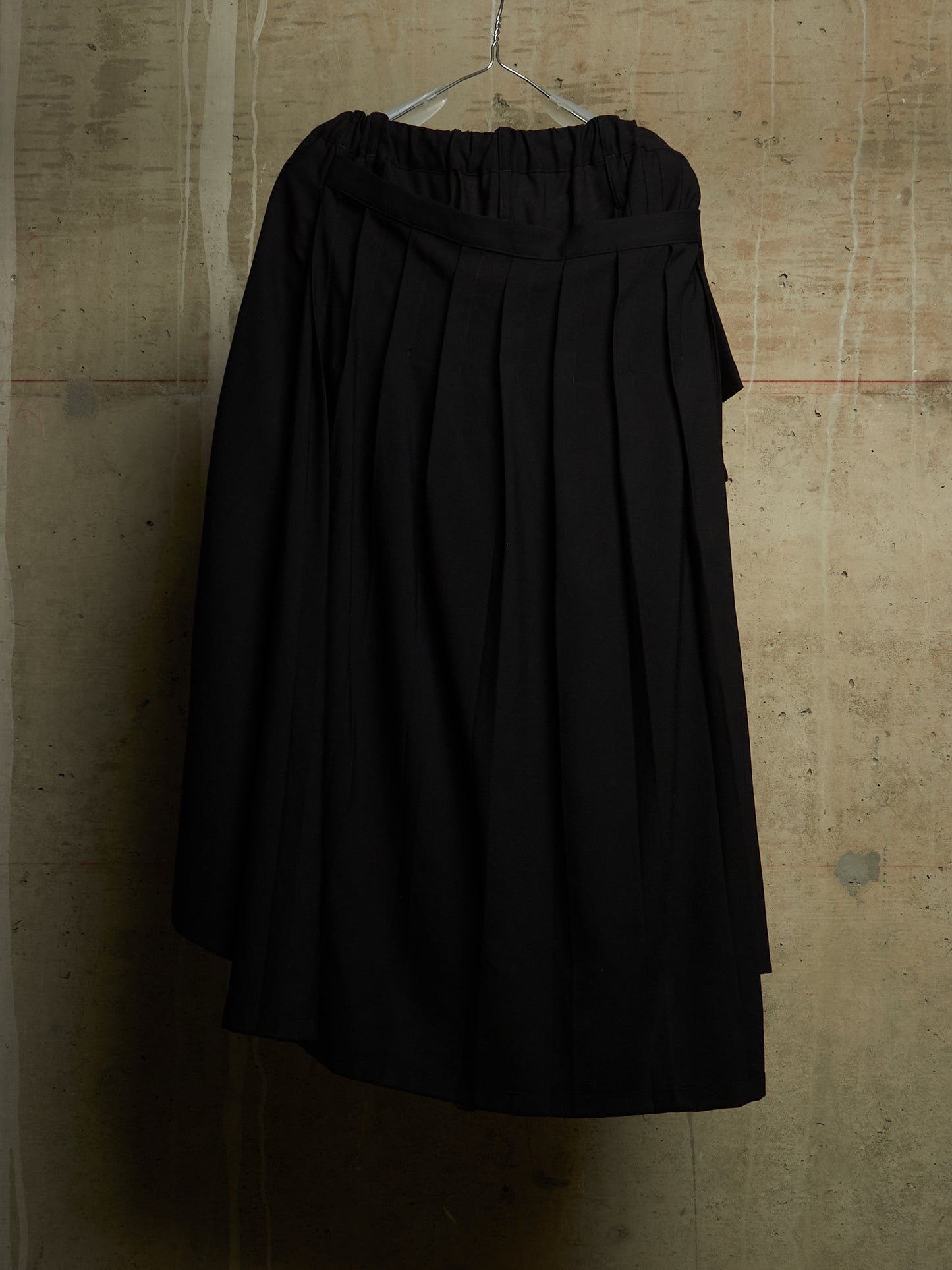 A black pleated midi skirt with an elastic waistband, hanging on a metal hanger against a textured concrete wall.