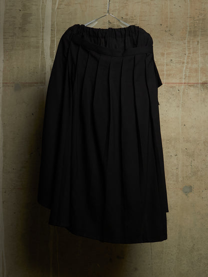 A black pleated midi skirt with an elastic waistband, hanging on a metal hanger against a textured concrete wall.