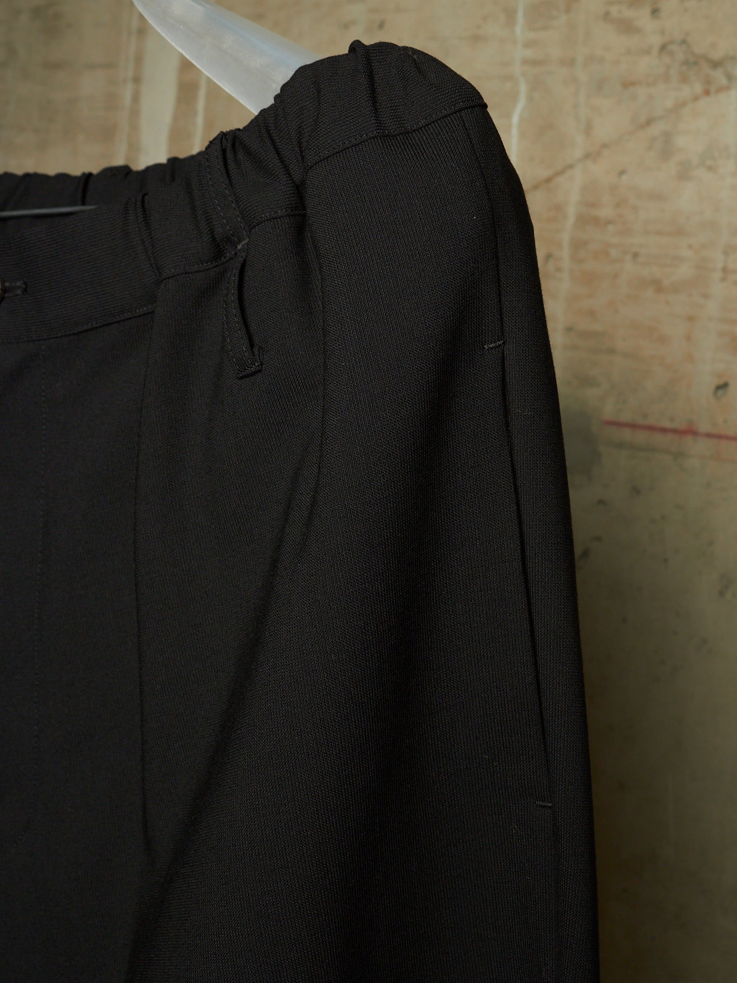 Close-up of black trousers hanging on a clear plastic hanger against a textured beige wall.