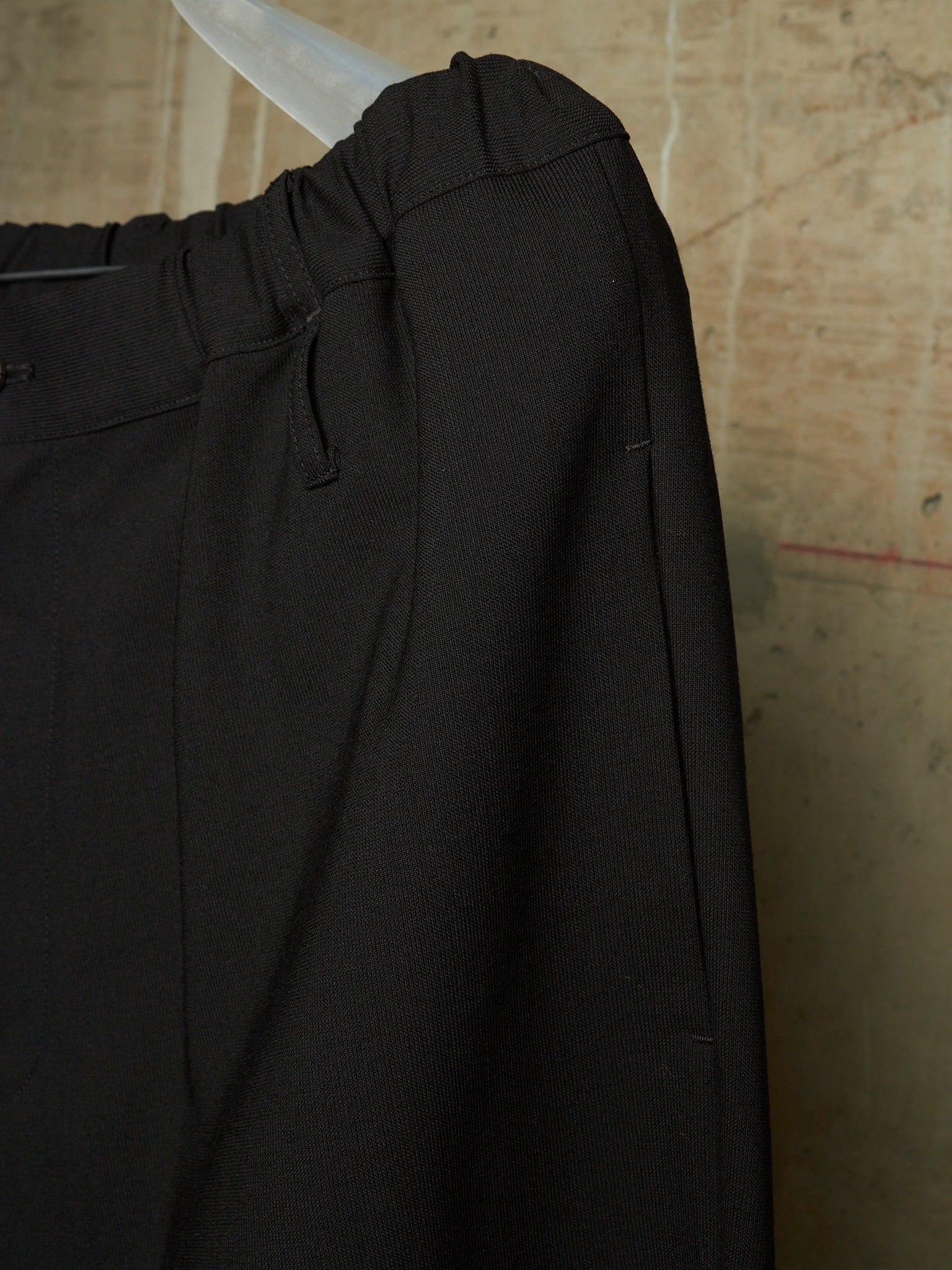 Close-up of black trousers hanging on a clear plastic hanger against a textured beige wall.