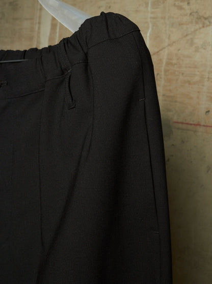 Close-up of black trousers hanging on a clear plastic hanger against a textured beige wall.