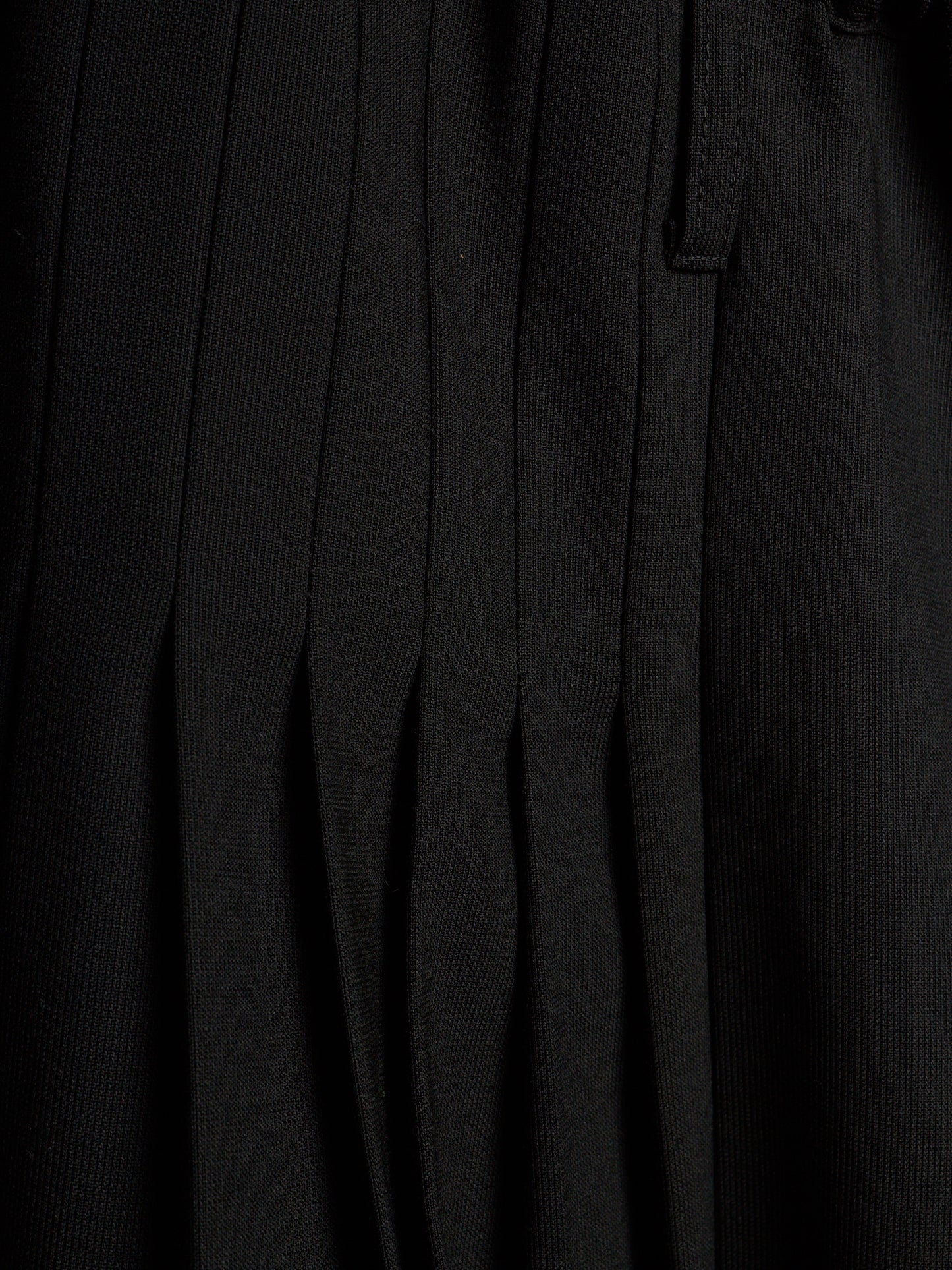 Close-up of a black pleated fabric garment, showing vertical pleats and a small tab detail.