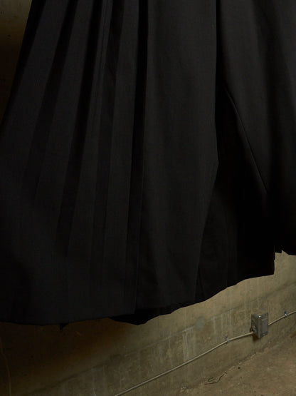 A close-up of a black pleated fabric garment, likely a skirt or drape, hanging against a textured concrete wall.