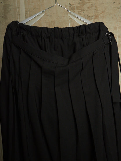 Black pleated trousers with a wide waistband and side tie details, hanging on a metal hanger against a textured beige wall.