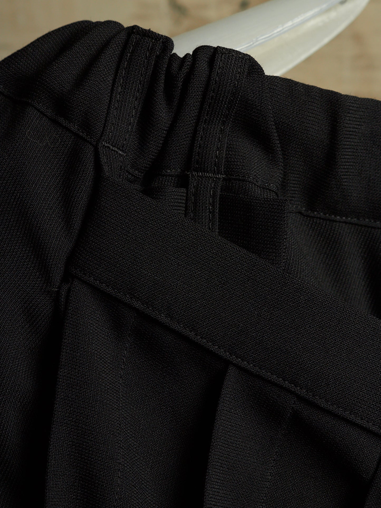 Close-up detail of black pleated fabric with a wide black belt crossing over the front, showing texture and stitching.