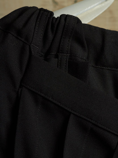 Close-up detail of black pleated fabric with a wide black belt crossing over the front, showing texture and stitching.