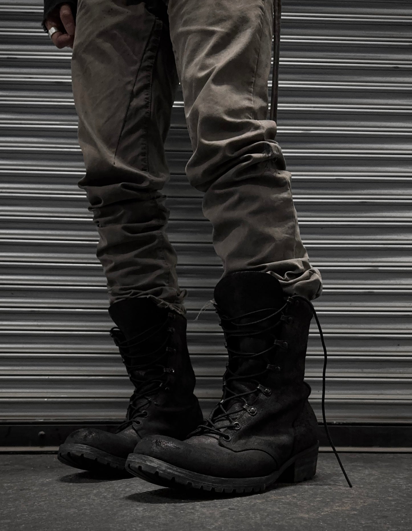 A person in gray pants and black ATTACHMENT by Kazuyuki Kumagai Reversed Cow Leather Lace-up Boots stands before a metallic, corrugated shutter. Only their legs and feet are visible, capturing the sleek style of Attachment menswear.