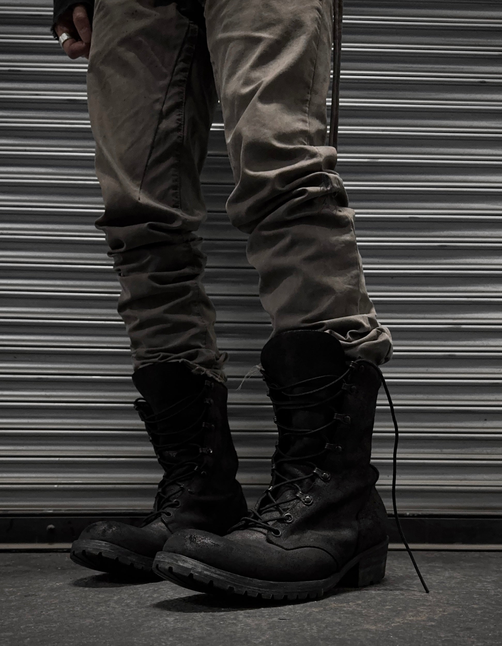 A person in gray pants and black ATTACHMENT by Kazuyuki Kumagai Reversed Cow Leather Lace-up Boots stands before a metallic, corrugated shutter. Only their legs and feet are visible, capturing the sleek style of Attachment menswear.