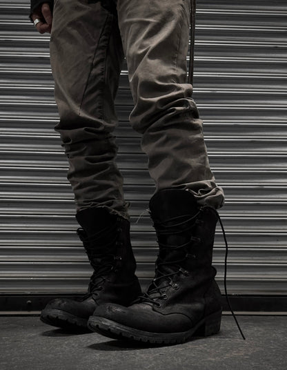 A person in gray pants and black ATTACHMENT by Kazuyuki Kumagai Reversed Cow Leather Lace-up Boots stands before a metallic, corrugated shutter. Only their legs and feet are visible, capturing the sleek style of Attachment menswear.