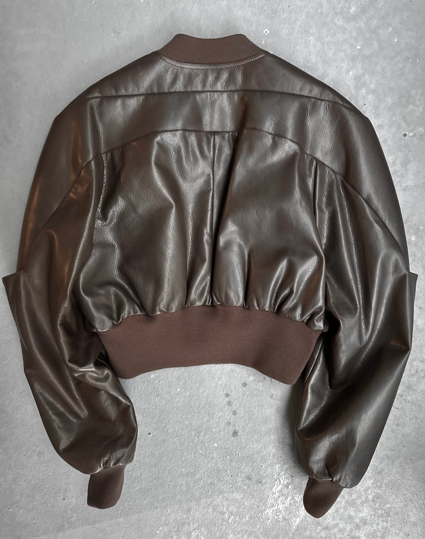 The Rick Owens AW23 LUXOR Girdered Leather Bomber Jacket, in dark brown calfskin, is displayed flat on a gray surface, back view visible, featuring ribbed knit cuffs, waistband and collar with a slightly oversized gathered silhouette.