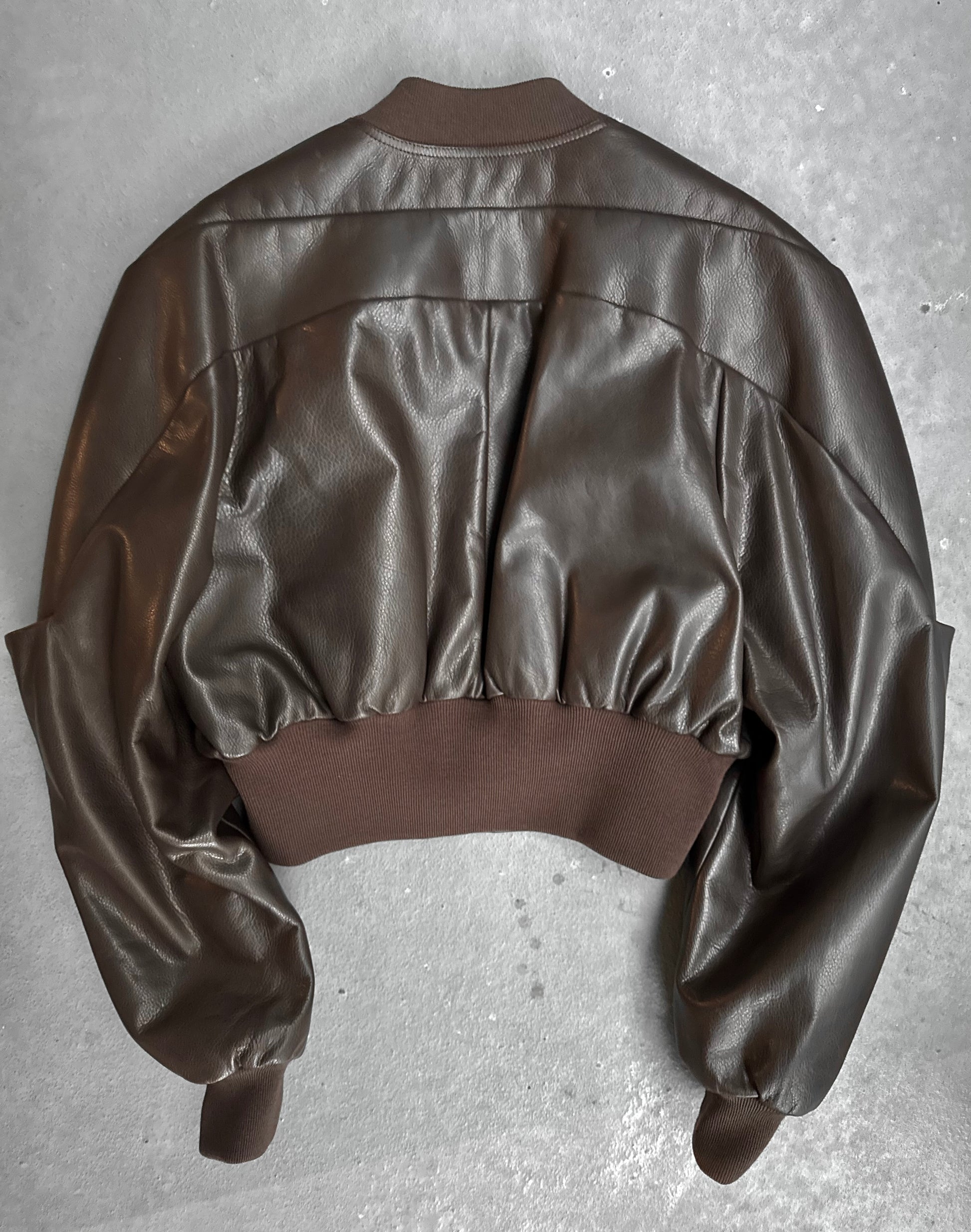 The Rick Owens AW23 LUXOR Girdered Leather Bomber Jacket, in dark brown calfskin, is displayed flat on a gray surface, back view visible, featuring ribbed knit cuffs, waistband and collar with a slightly oversized gathered silhouette.