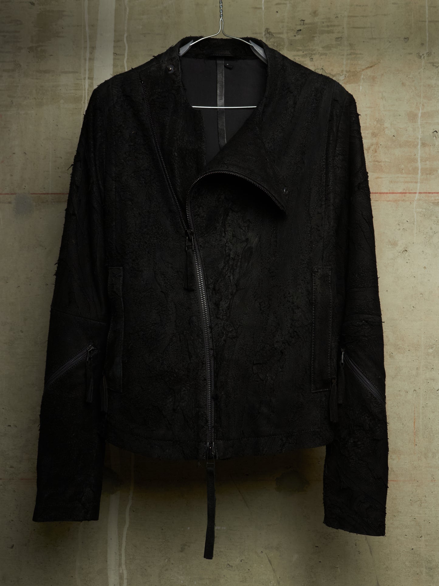 A black textured moto jacket with a distressed, frayed edge and asymmetrical zipper, hanging against a concrete wall.