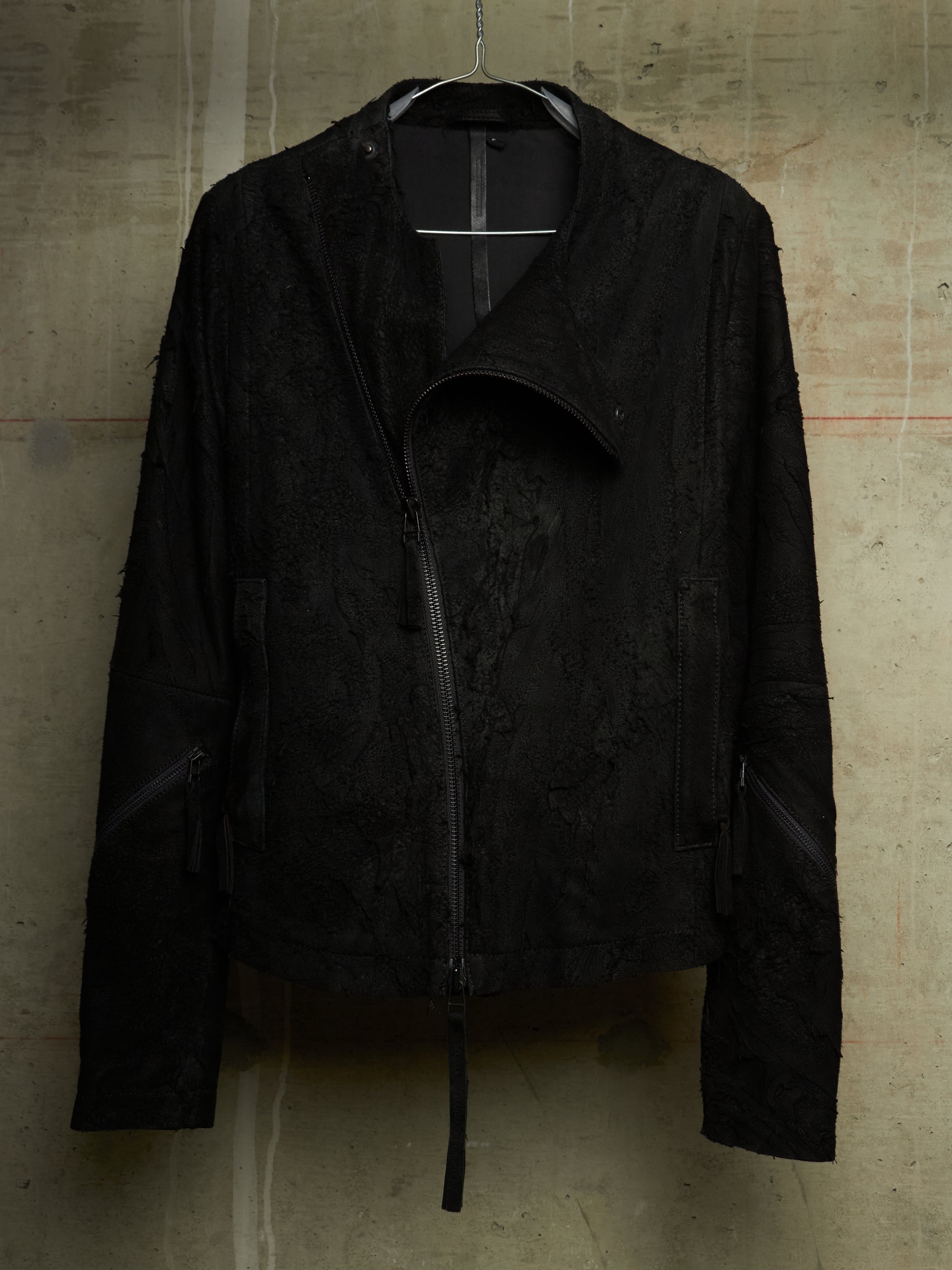 A black textured moto jacket with a distressed, frayed edge and asymmetrical zipper, hanging against a concrete wall.