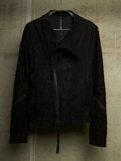 A black textured moto jacket with a distressed, frayed edge and asymmetrical zipper, hanging against a concrete wall.