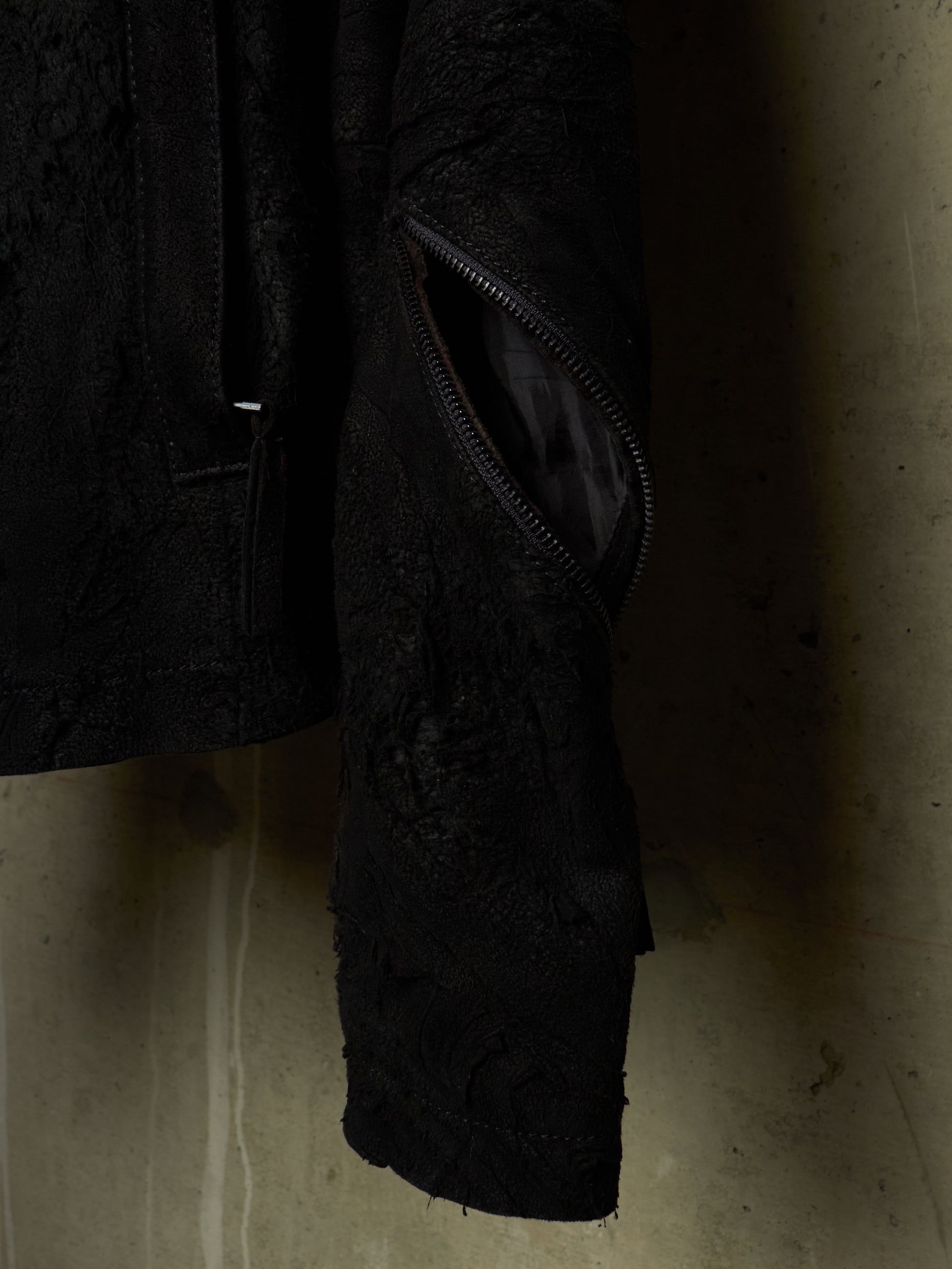 Close-up of a black textured jacket with a diagonal zippered pocket, hanging against a concrete wall.