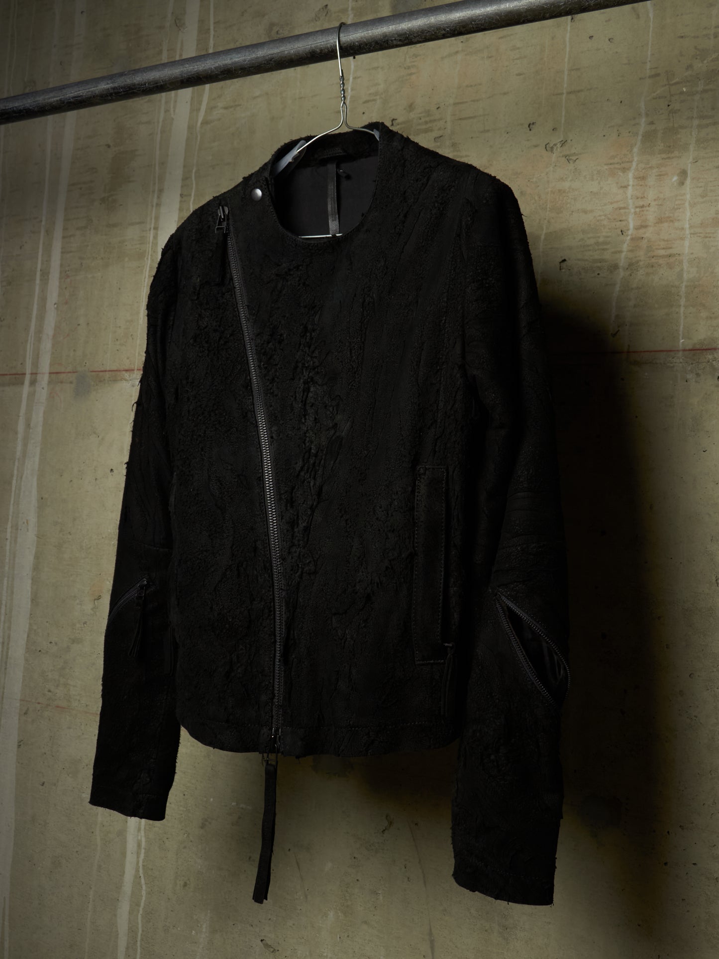 A black textured jacket with a diagonal zipper, hanging on a metal hanger from a horizontal rod against a concrete wall.