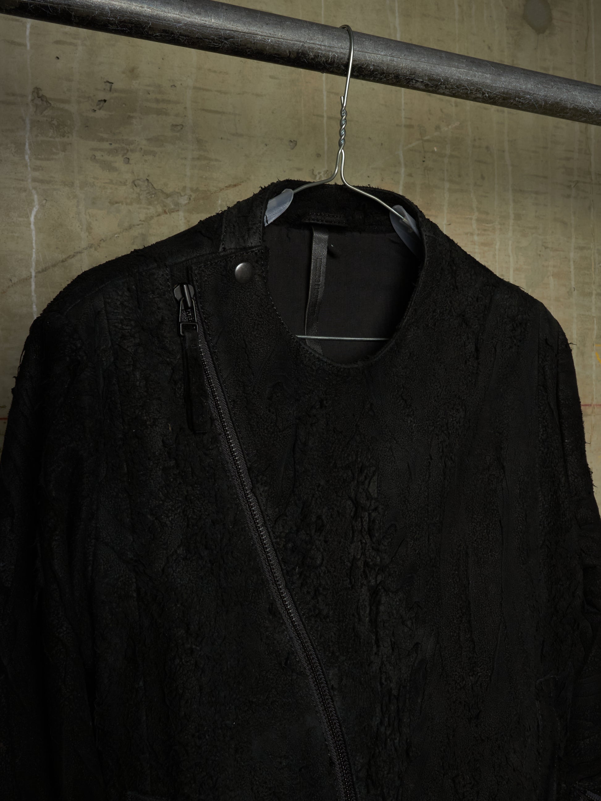 A black textured jacket with a diagonal zipper, hanging on a black hanger from a metal pipe against a concrete wall.