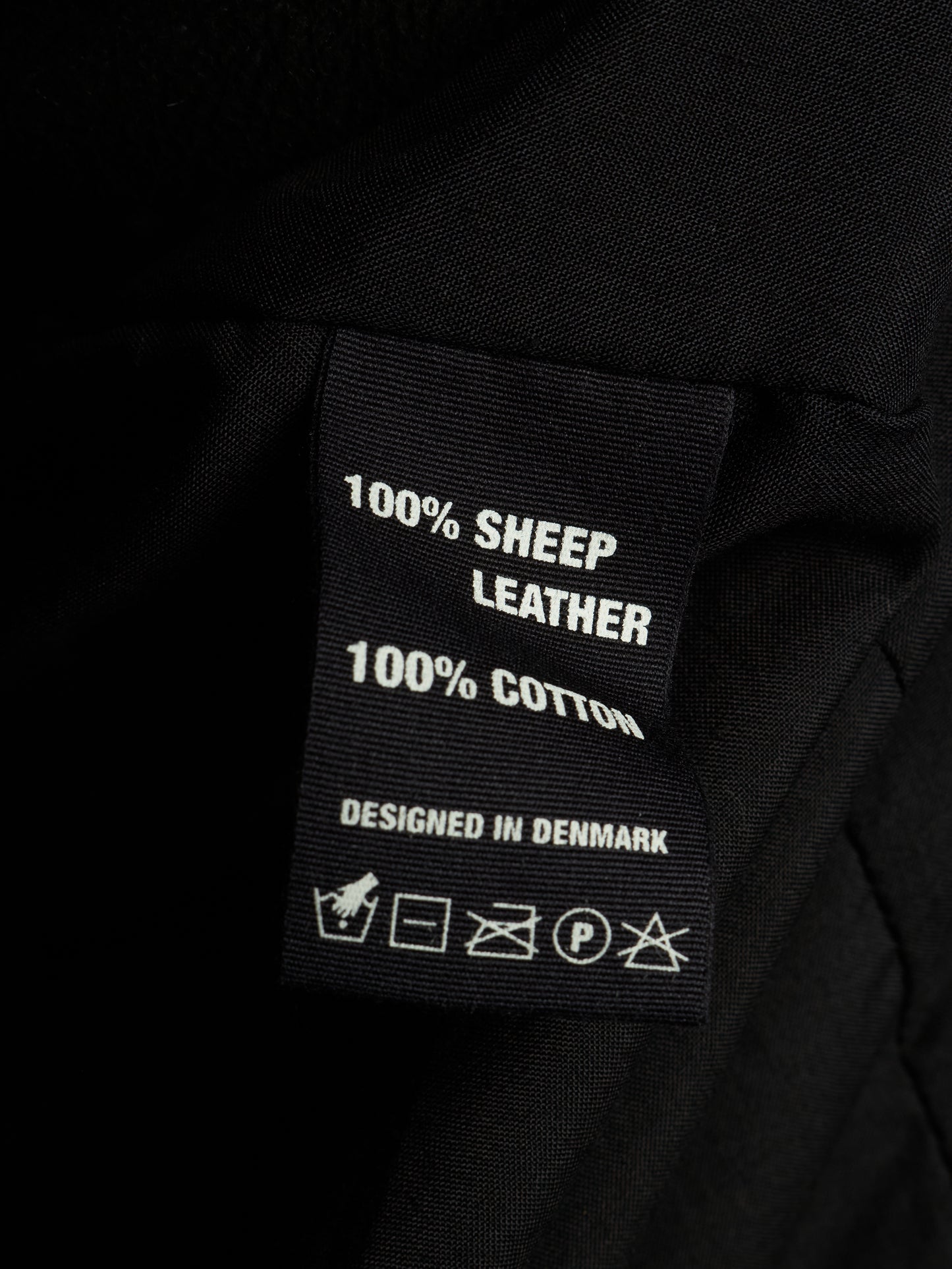 Close-up of a black fabric garment label reading '100% SHEEP LEATHER' and '100% COTTON' with care symbols below, set against dark black fabric.