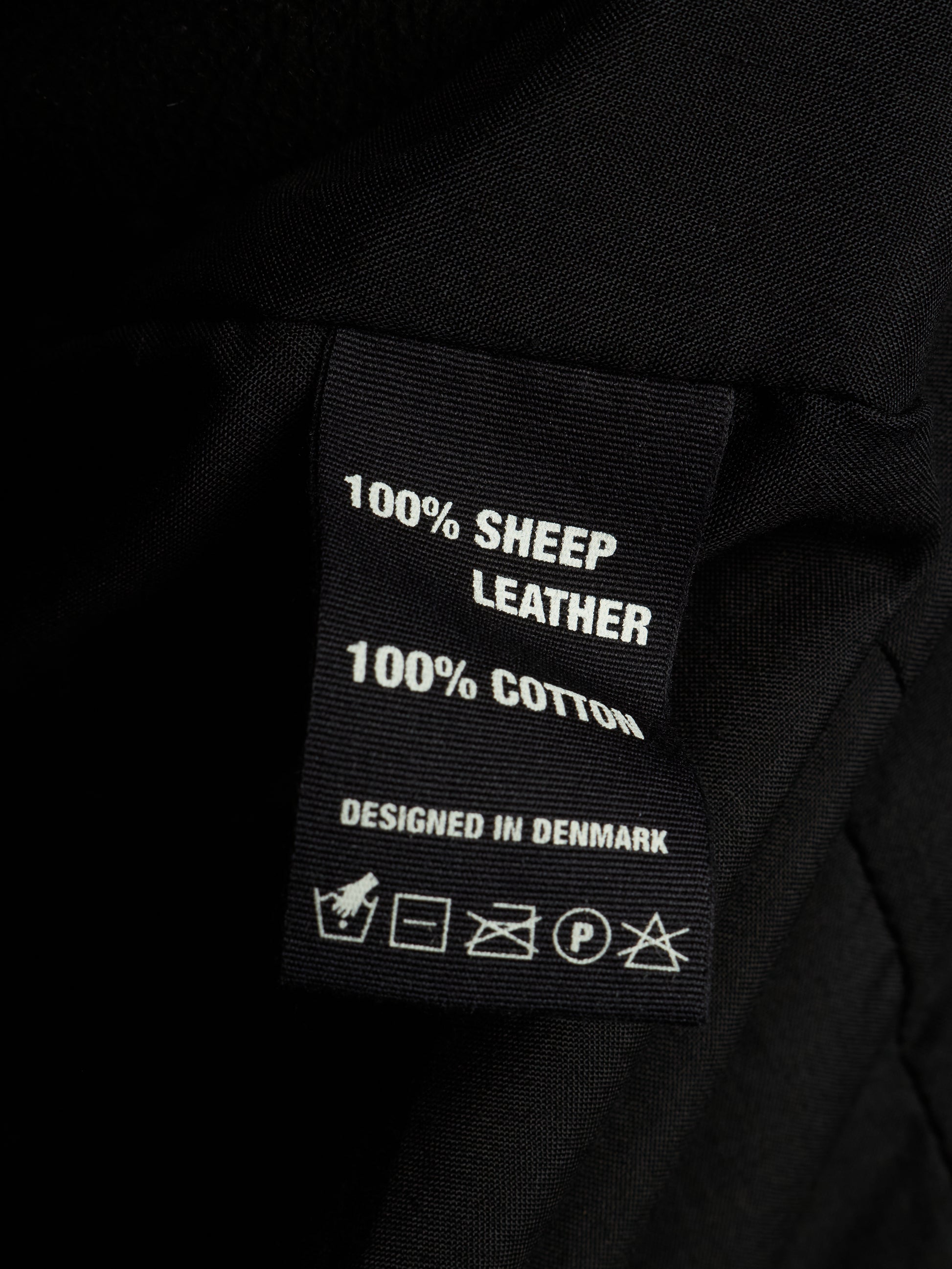 Close-up of a black fabric garment label reading '100% SHEEP LEATHER' and '100% COTTON' with care symbols below, set against dark black fabric.