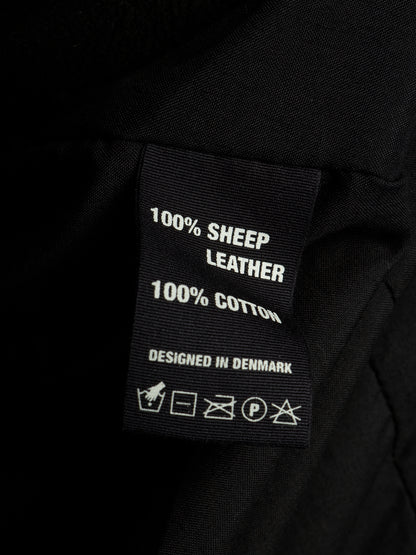 Close-up of a black fabric garment label reading '100% SHEEP LEATHER' and '100% COTTON' with care symbols below, set against dark black fabric.