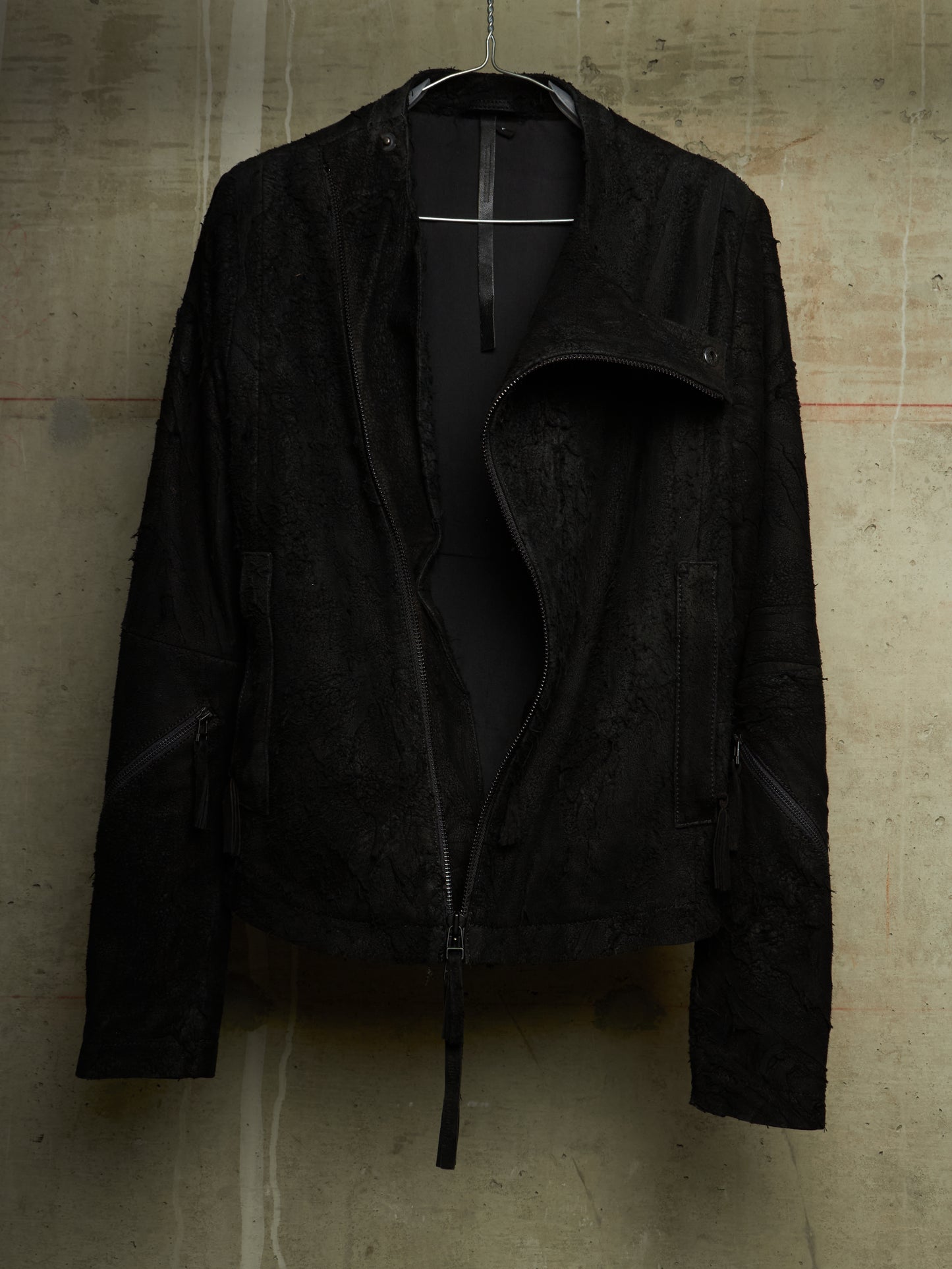 A black textured moto jacket with an asymmetrical zipper and side zippers, hanging on a black hanger against a concrete wall.