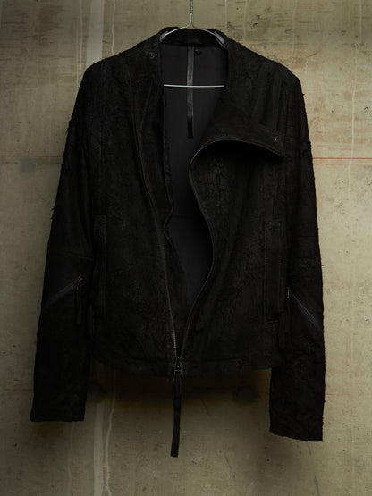 A black textured moto jacket with an asymmetrical zipper and side zippers, hanging on a black hanger against a concrete wall.