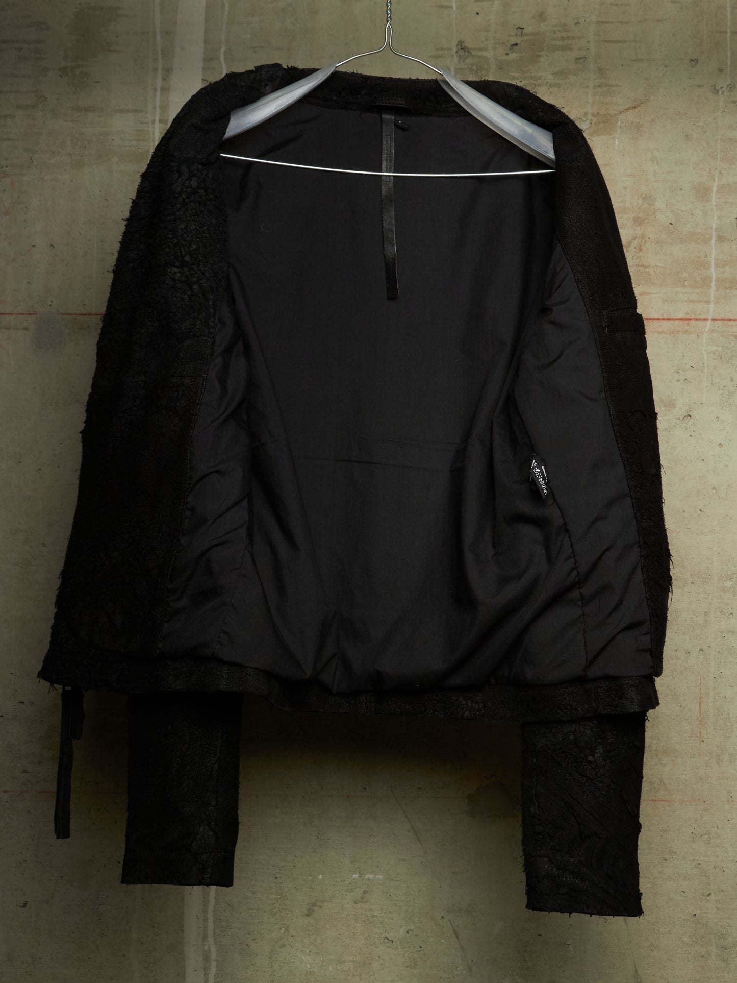 A black cropped jacket with textured fuzzy sleeves and a zippered front, hanging on a metal hanger against a textured concrete wall.